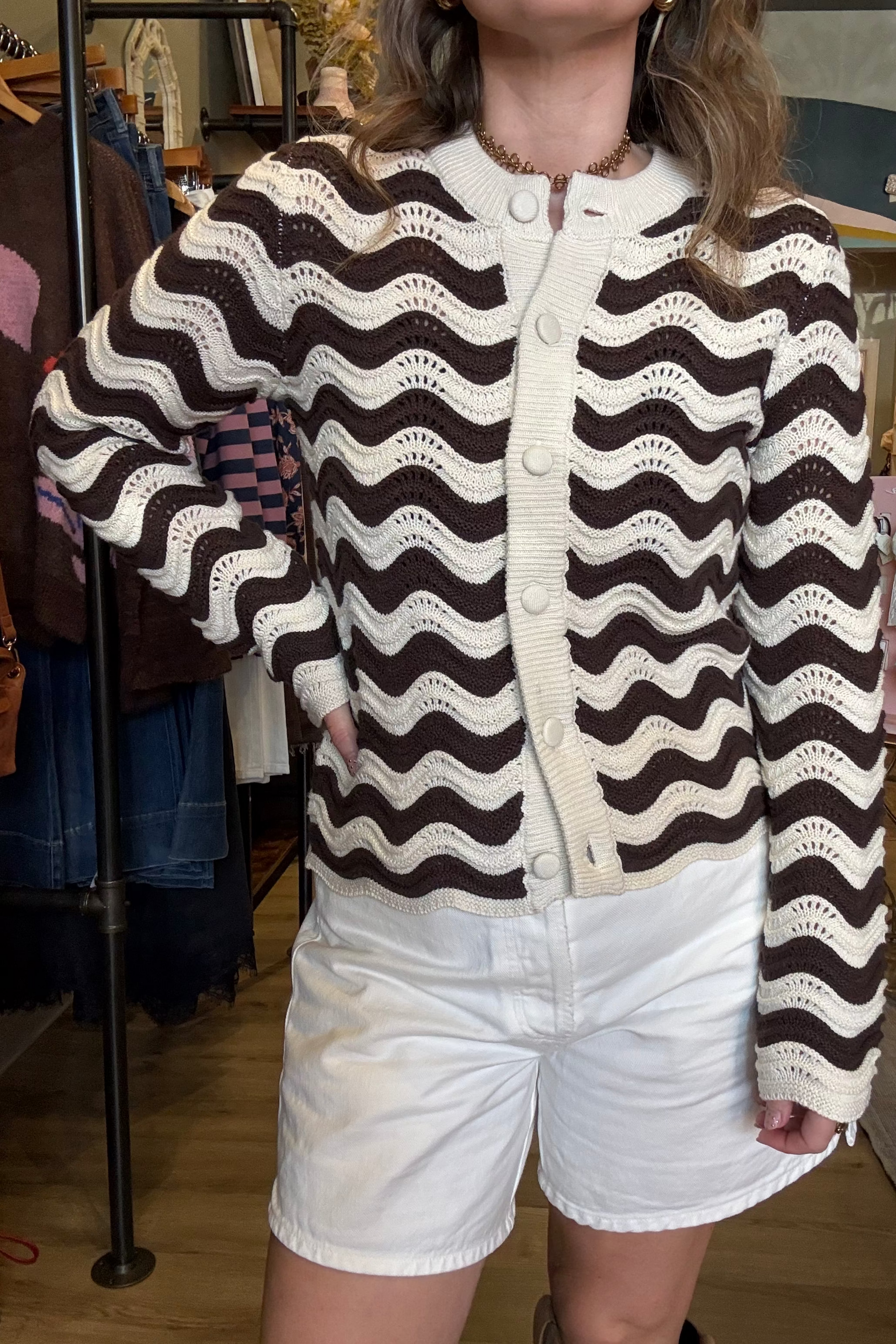 A person wearing a brown and cream wavy knit cardigan with white shorts, standing inside a clothing store.