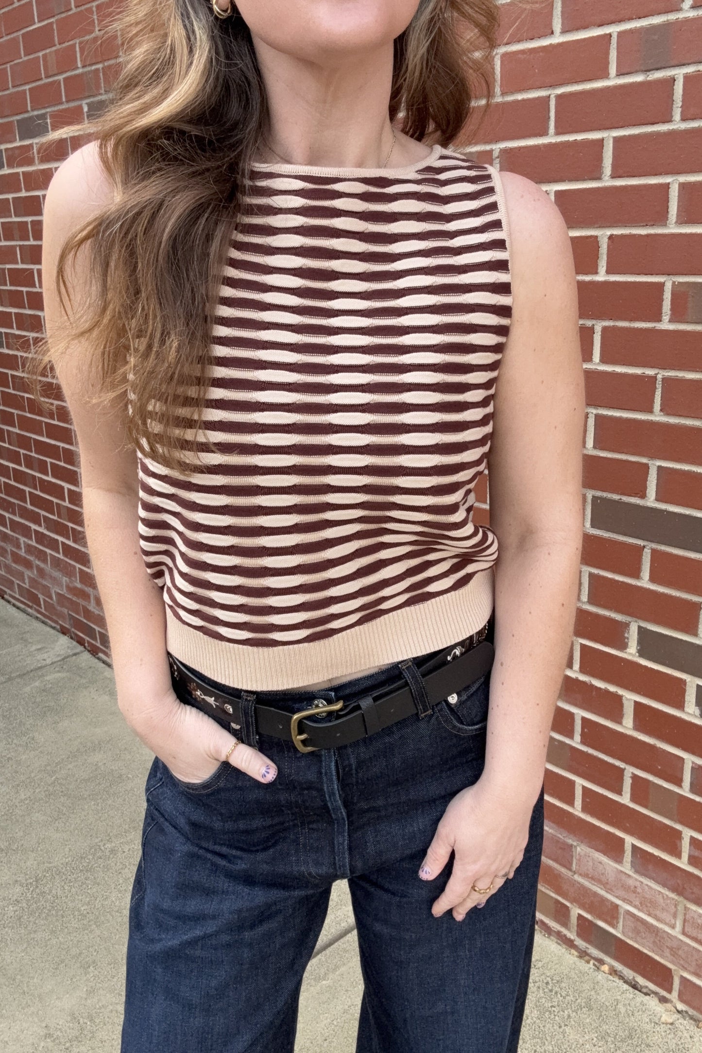 Person wearing a brown and beige striped sleeveless top and blue jeans against a brick wall.