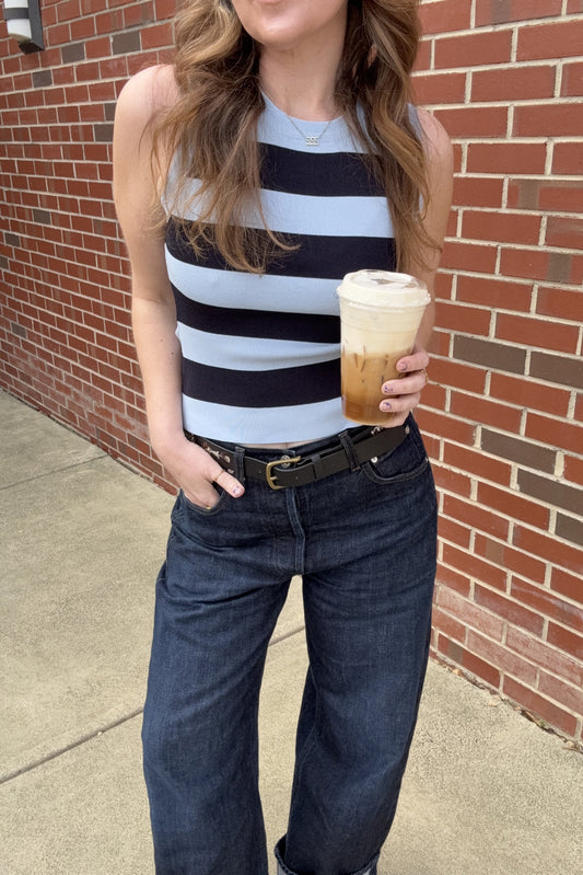 Woman in a striped tank top and jeans holding a coffee cup against a brick wall.