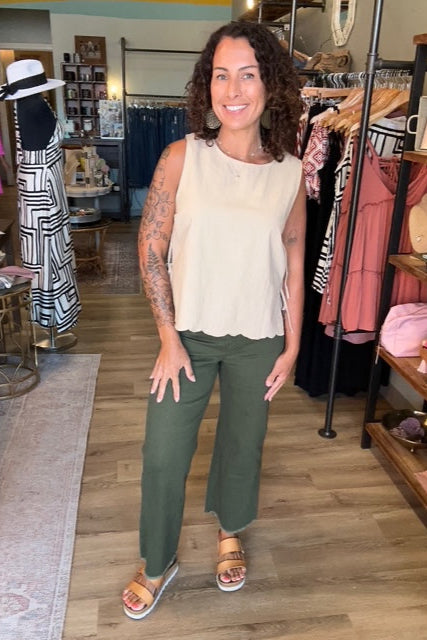 Woman wearing a white top and green pants standing in a store with racks and products in the background.