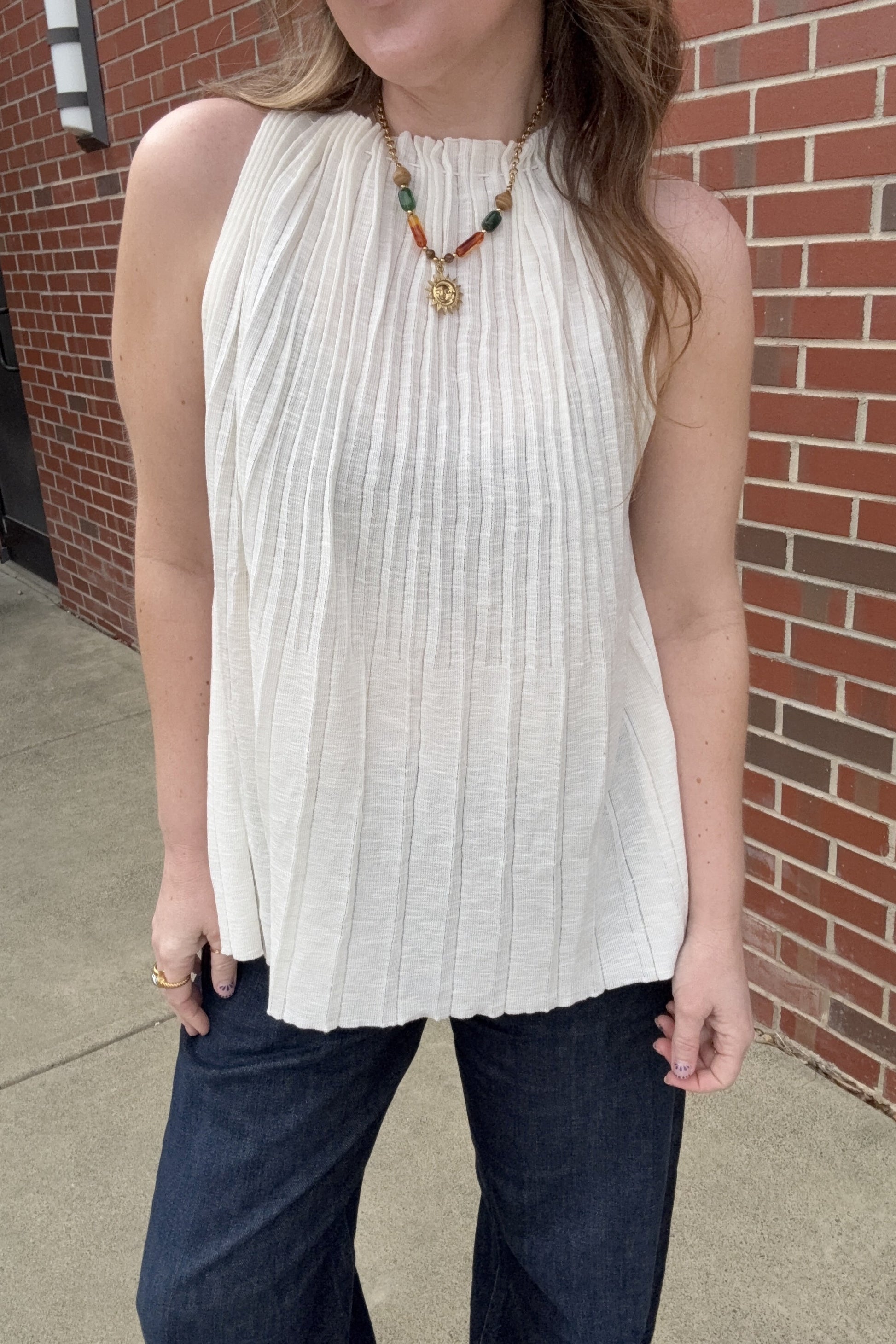 Person wearing a white sleeveless top and dark pants in front of a brick wall.