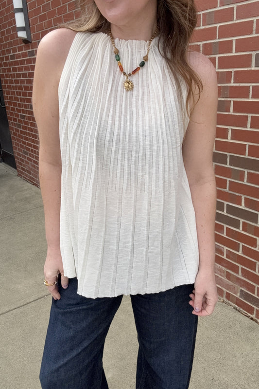Person wearing a white sleeveless top and dark pants in front of a brick wall.