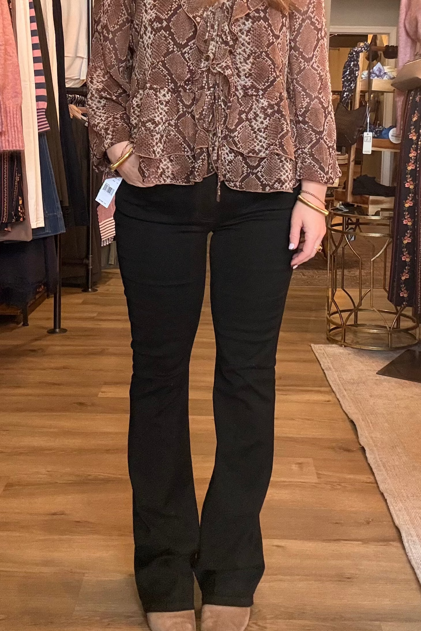 A person standing in a clothing store wearing a brown and beige snakeskin-patterned long-sleeve top and black flare pants, paired with tan boots.