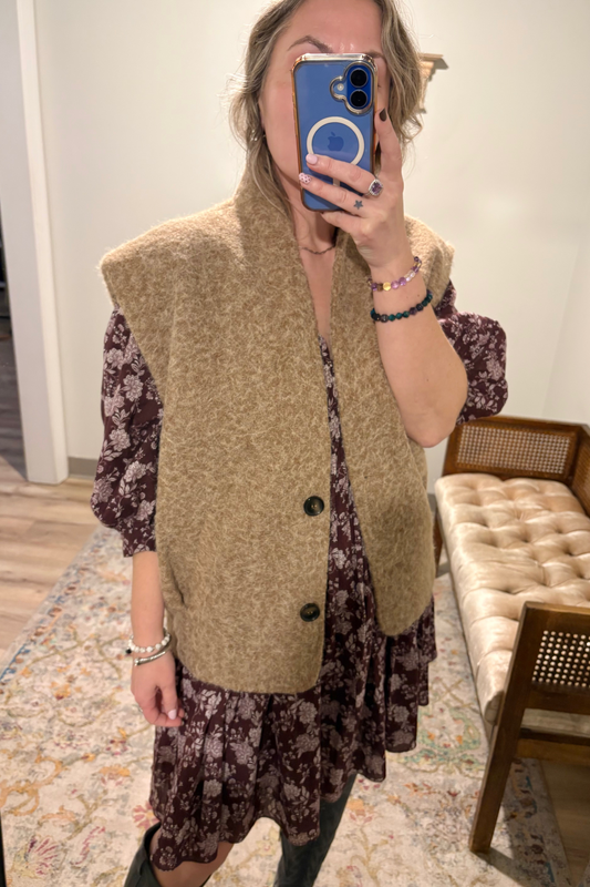 A woman takes a mirror selfie in a boutique showing a two-piece outfit consisting of a chunky, textured tan sleeveless vest and a dark brown floral print dress with puff sleeves.