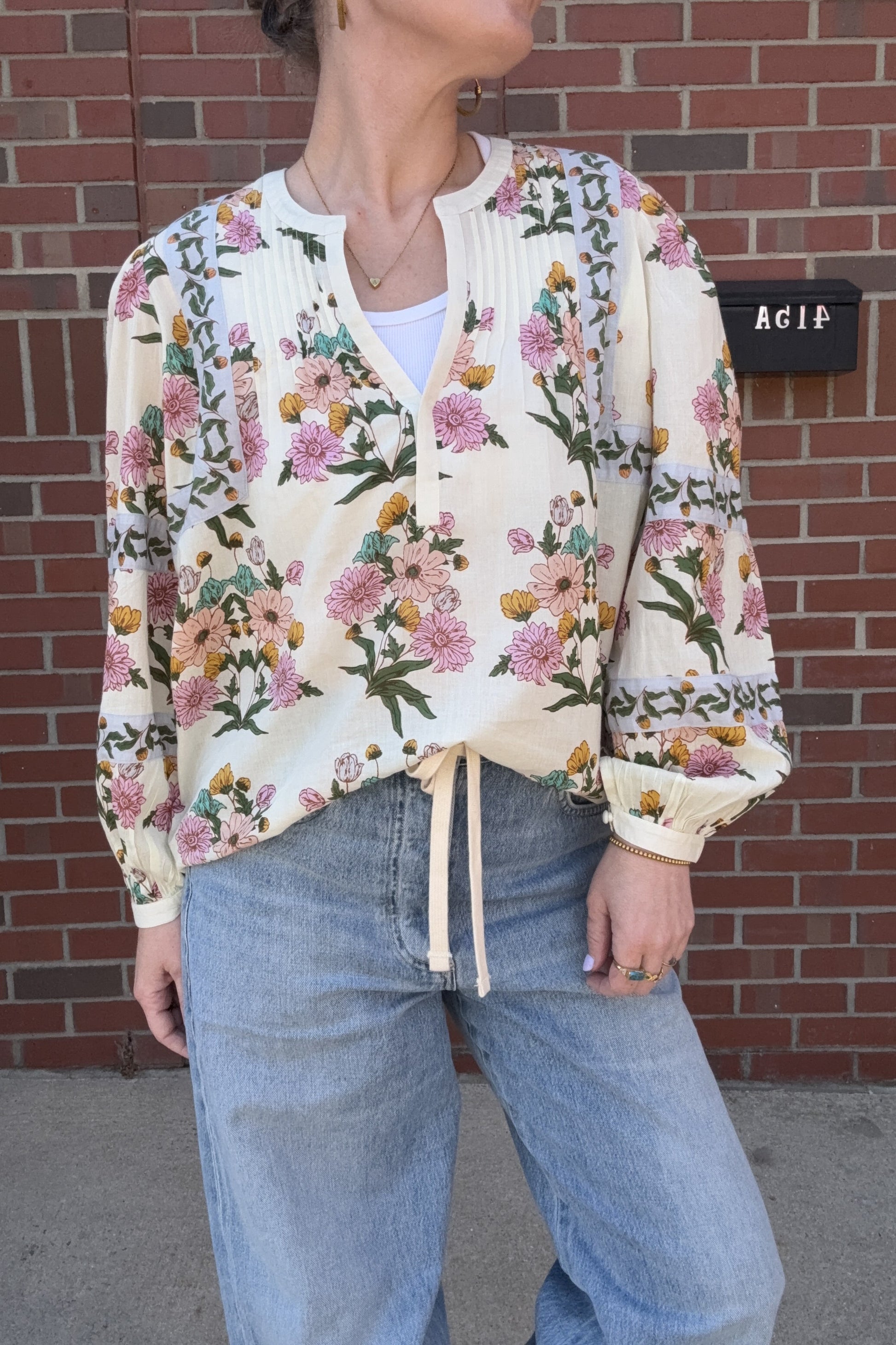 Person wearing a floral blouse and jeans against a brick wall.