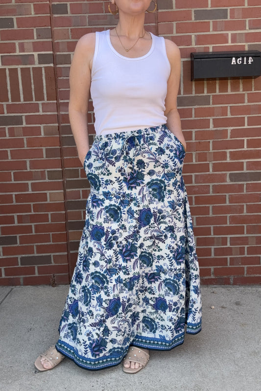 Person wearing a white tank top and blue floral skirt standing in front of a brick wall.