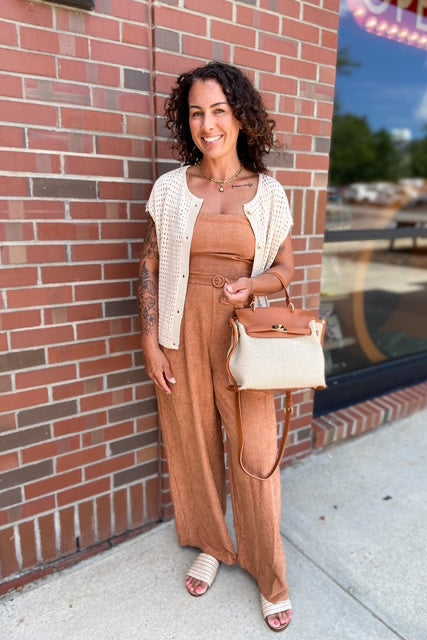 Woman in a brown jumpsuit standing against a brick wall with a store window in the background.