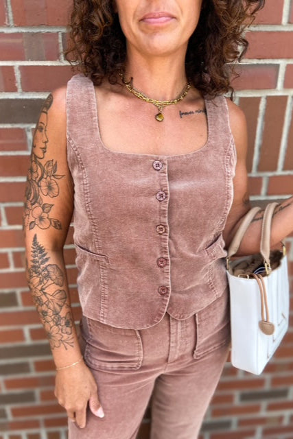Woman wearing a brown corduroy outfit with tattoos visible on arms, standing against a brick wall.