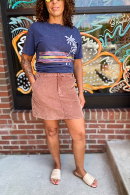 Person wearing a blue t-shirt with a colorful design and a brown skirt standing in front of a colorful mural.