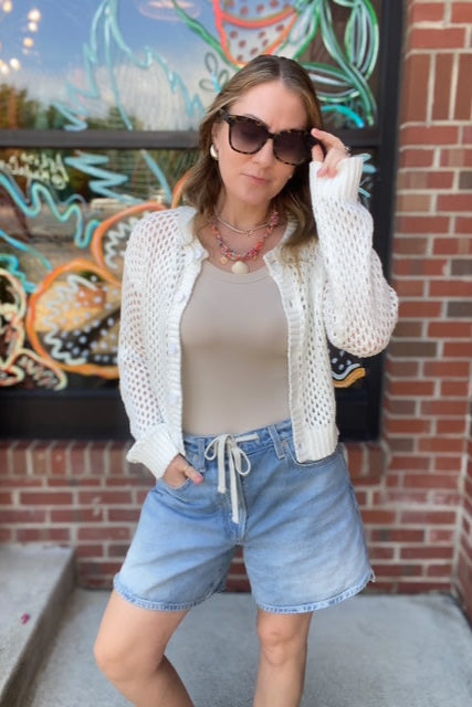 Woman wearing a white cardigan, beige top, and denim shorts in front of a brick wall with a colorful mural.
