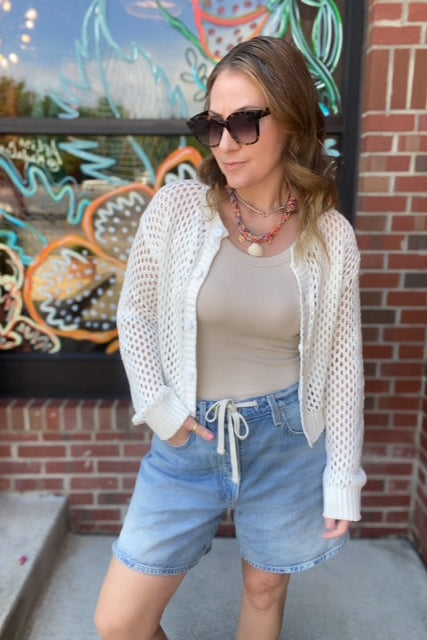 Woman wearing a white cardigan, beige top, and denim shorts standing in front of a colorful mural.