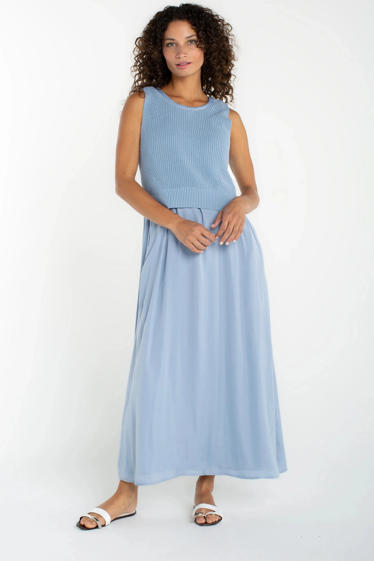 A woman modeling a sleeveless light blue maxi dress with a ribbed textured bodice and a flowing skirt, paired with white slide sandals, against a plain white studio background.
