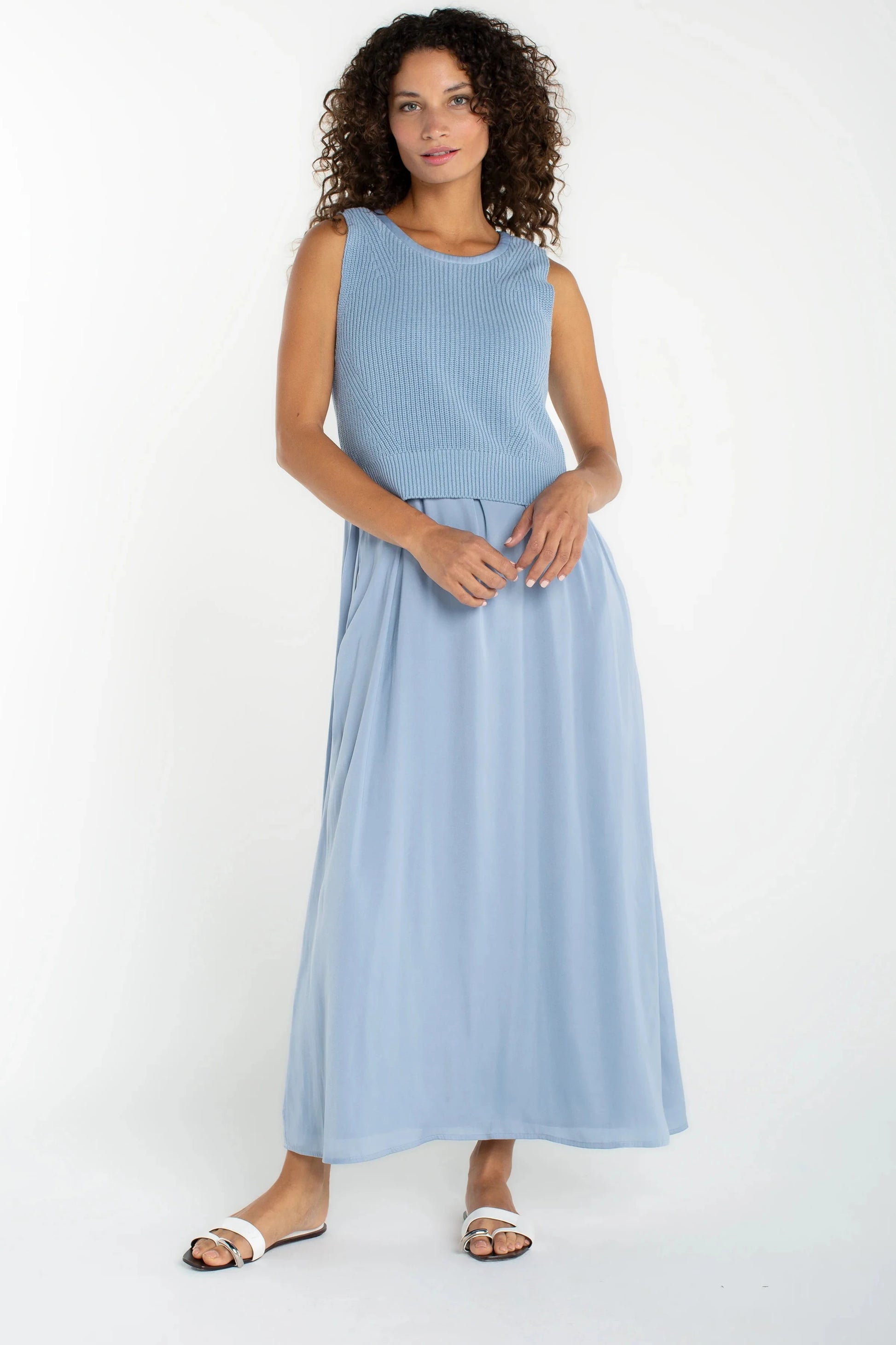 A woman modeling a sleeveless light blue maxi dress with a ribbed textured bodice and a flowing skirt, paired with white slide sandals, against a plain white studio background.