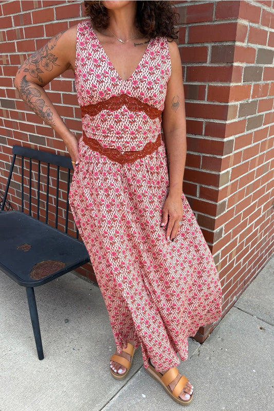 A woman wearing a sleeveless bohemian-style maxi dress with a red, pink, and white floral pattern and a brown lace waist detail, standing outdoors next to a brick wall.