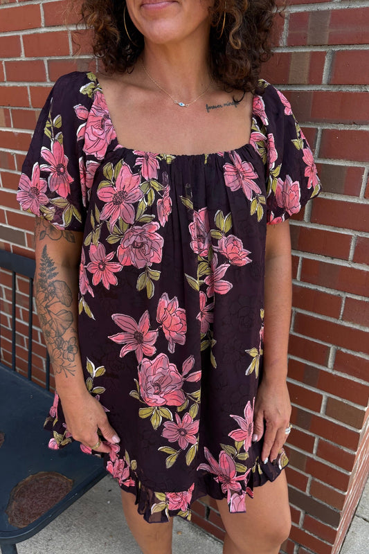 A woman wearing a dark purple short-sleeve dress with a pink floral print, standing in front of a red brick wall.