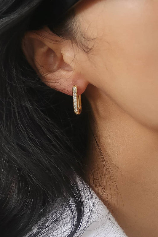 A close-up of a model's ear wearing a small gold huggie hoop earring adorned with clear pavé stones.