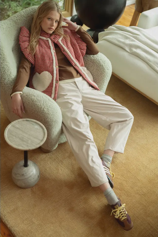 Woman sitting on a plush chair wearing a red checkered vest and white pants in a cozy room.