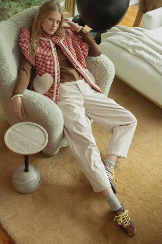 Woman sitting on a plush chair wearing a red checkered vest and white pants in a cozy room.