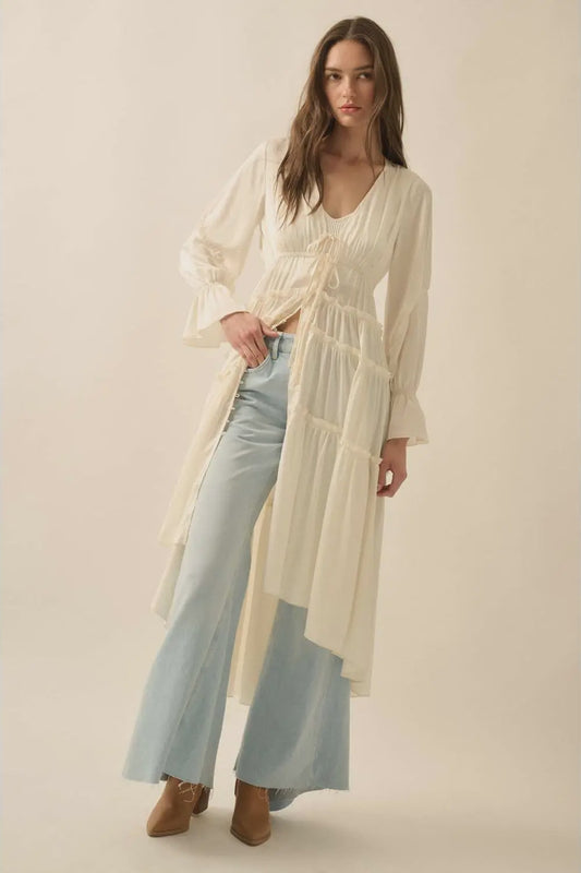 Woman wearing a long, light-colored dress over light blue jeans on a plain background