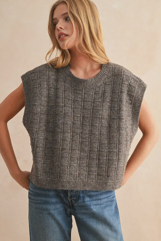 Woman wearing a gray sleeveless knitted top and blue jeans against a beige background