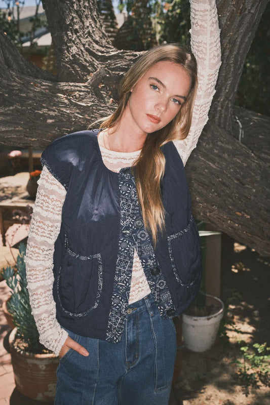 Woman wearing a blue vest over a white sweater, standing outdoors near a tree.