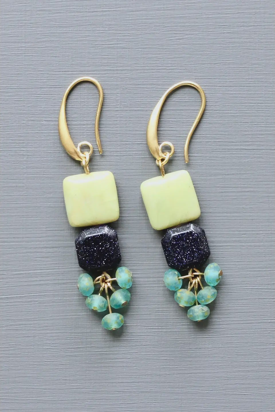 Pair of earrings with gold hooks, light green square beads, dark blue cylindrical beads, and green bead strings on a gray background.