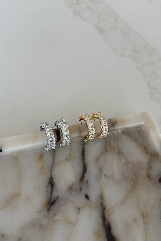 Two small huggie hoop earrings, one silver-toned and one gold-toned, both paved with clear stones, displayed on a white marble tray.