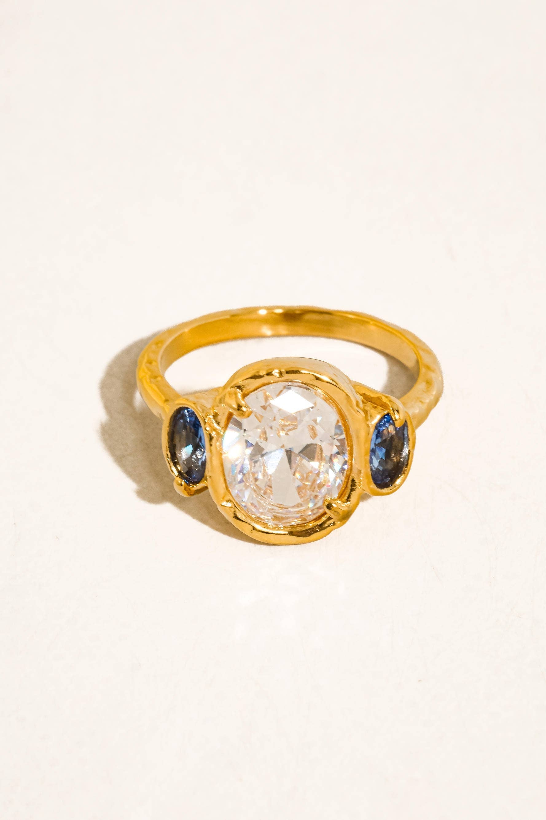 A gold ring featuring a large oval clear center stone flanked by two small round blue stones, photographed on a plain light background.