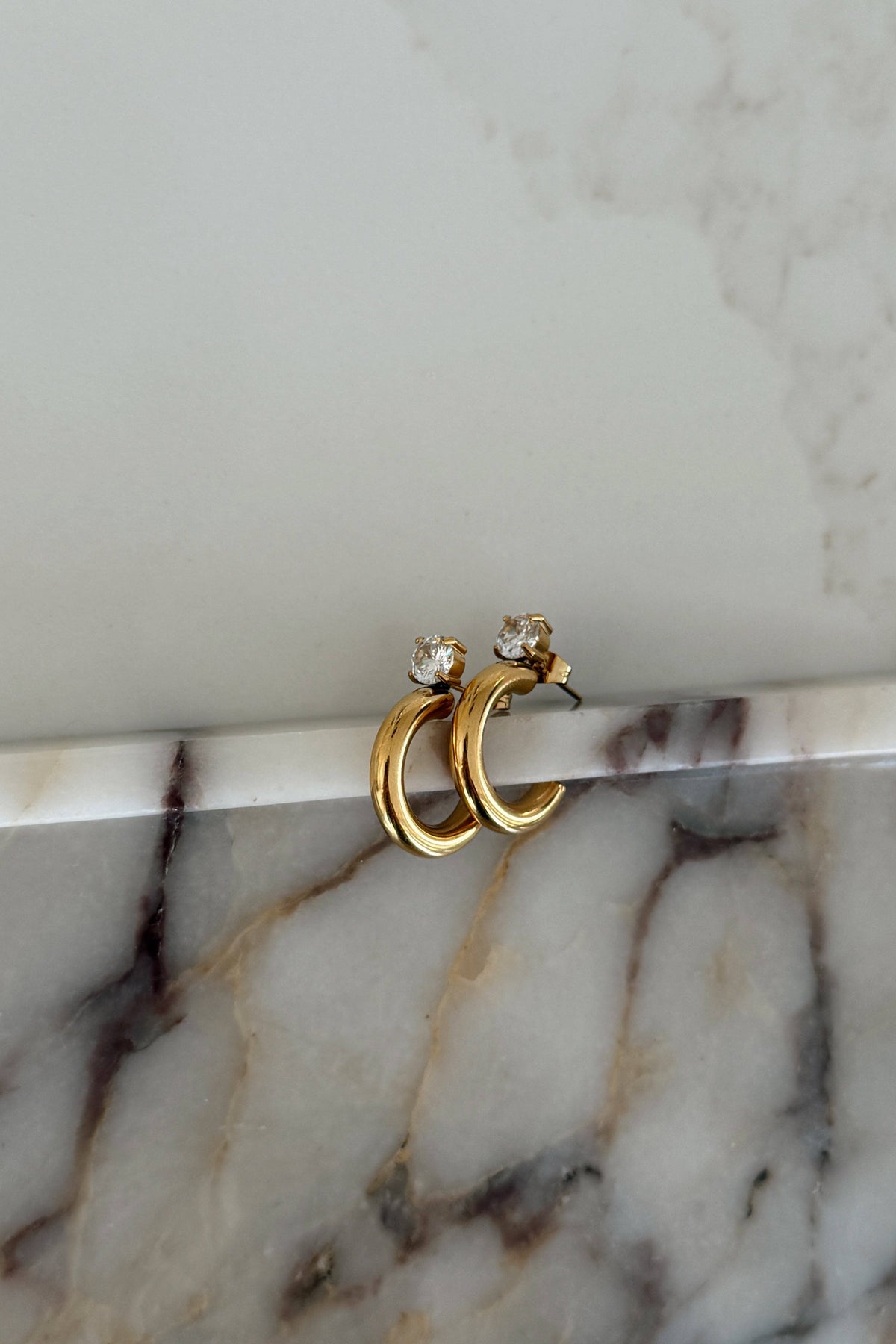 A pair of gold huggie hoop earrings with small clear gemstone accents, resting on a white marble surface against a light gray marble background.