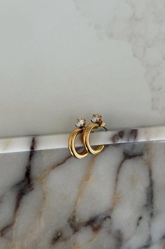 A pair of gold huggie hoop earrings with small clear gemstone accents, resting on a white marble surface against a light gray marble background.