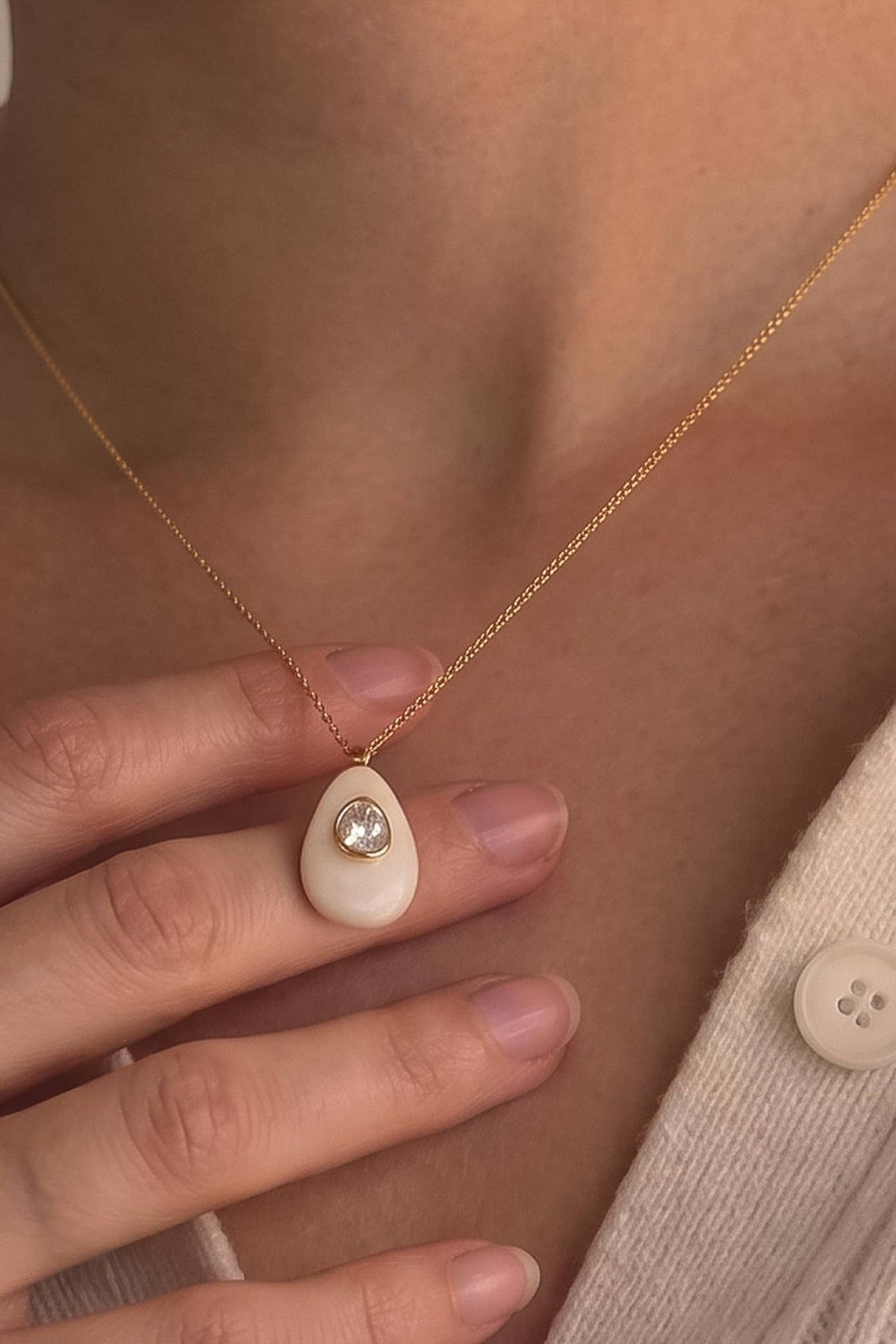 A close-up of a person wearing a delicate gold chain necklace with a small white teardrop-shaped pendant featuring a central clear gemstone, styled with a cream-colored knit top.