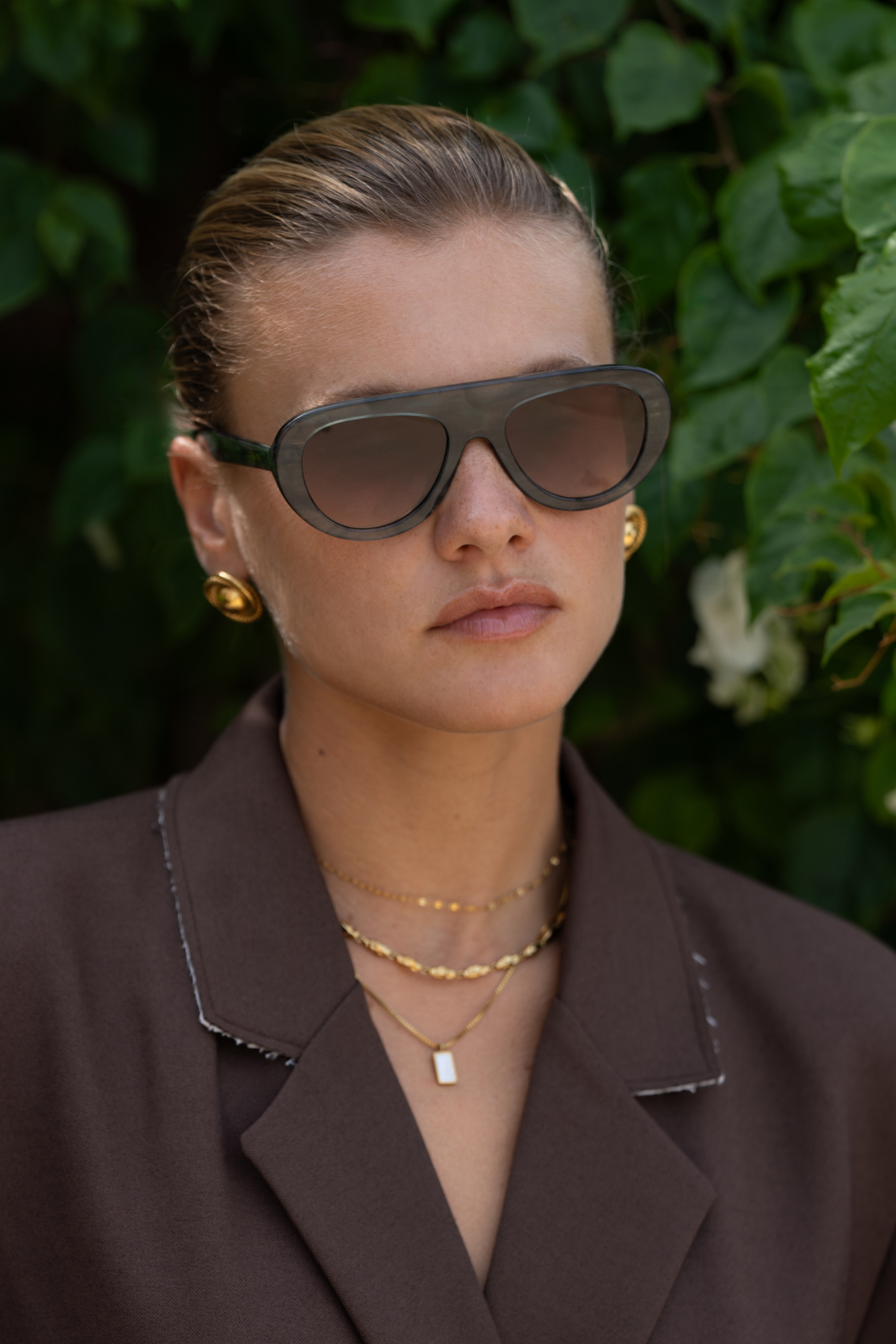 Woman wearing sunglasses and a brown outfit with a green leafy background