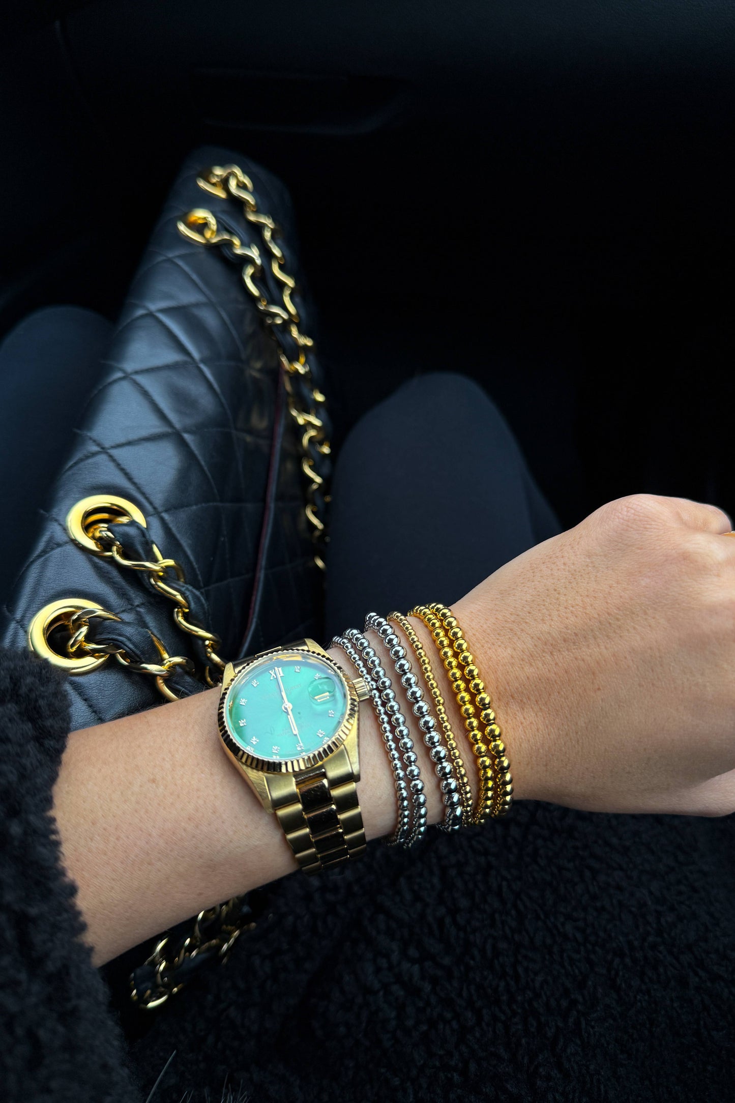 Hand wearing a gold watch and bracelets with a black quilted handbag in the background