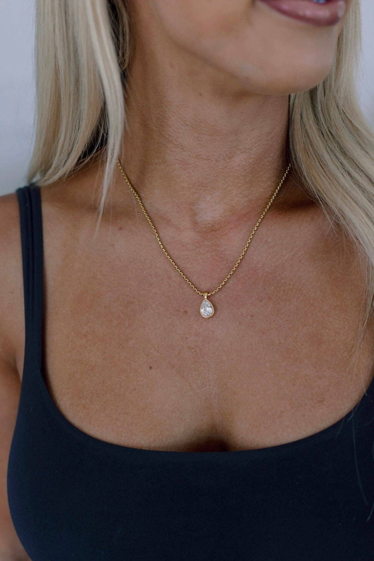 A close-up of a woman wearing a delicate gold chain necklace with a teardrop-shaped white pearl pendant, paired with a black tank top.