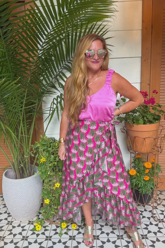A woman modeling a two-piece outfit consisting of a bright pink ruffled sleeveless top and a matching high-low midi skirt with a pink and gray tropical floral print, styled with gold metallic sandals and sunglasses in an outdoor patio setting with potted plants.