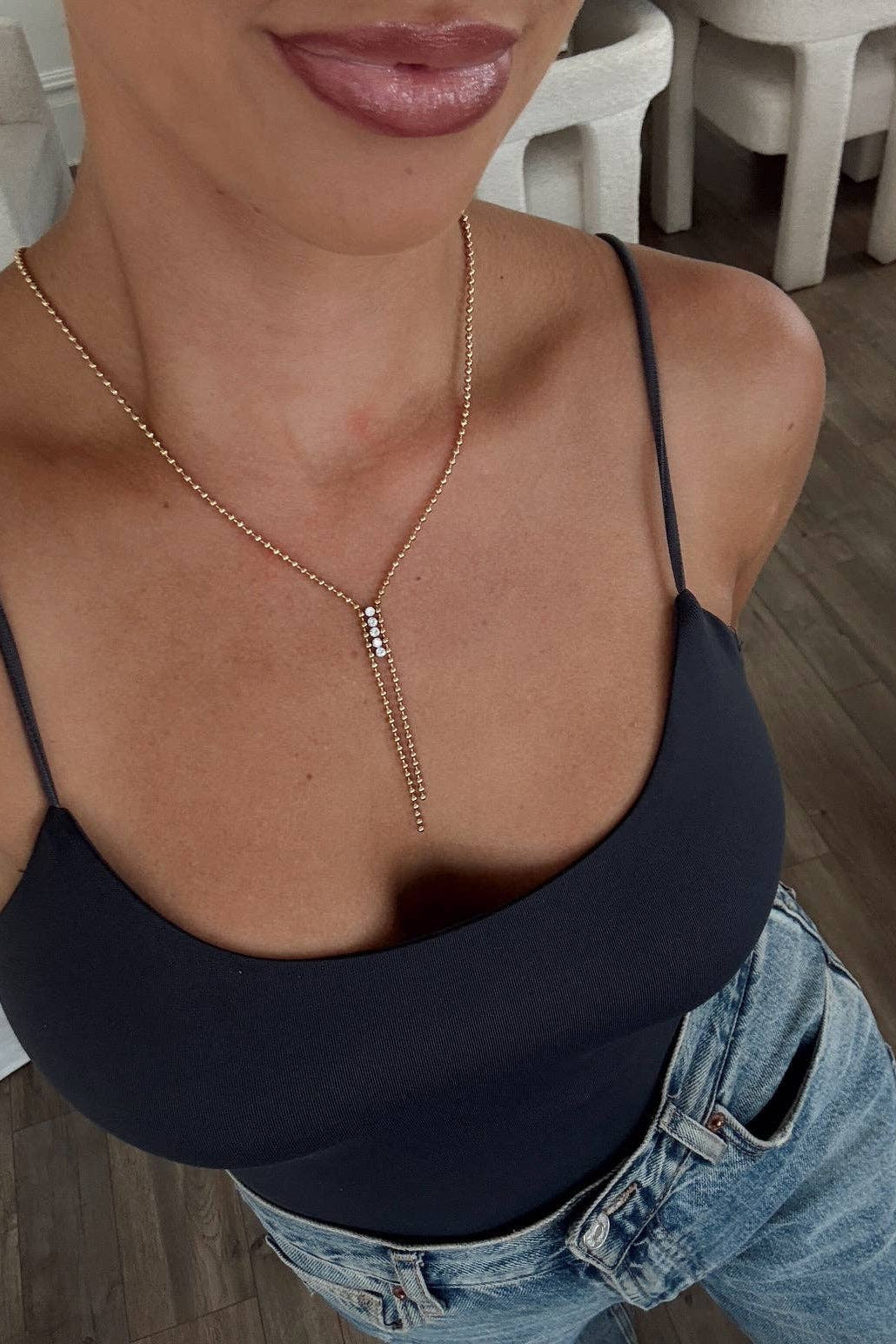 A close-up of a woman wearing a delicate gold Y-necklace with a central silver-toned bar detail, styled with a black camisole top and blue jeans.