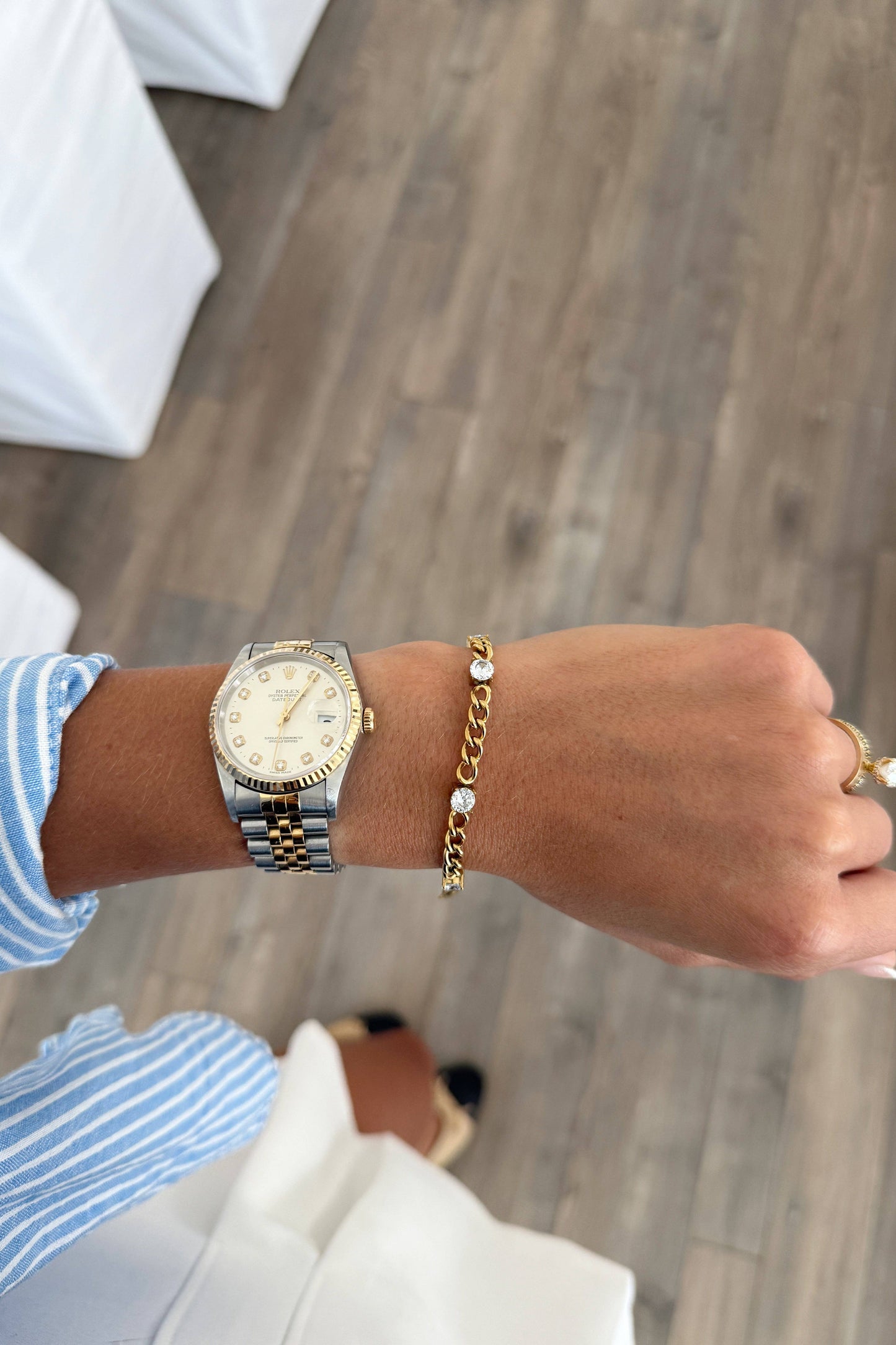 A person's wrist wearing a gold and silver two-tone watch and a gold chain bracelet with a pearl accent, shown against a background of a gray wood floor and white draped tables.