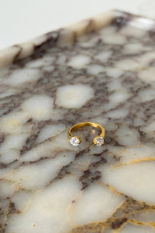 A gold open-bend ring with two small clear gemstones, displayed on a polished white and brown marble slab.