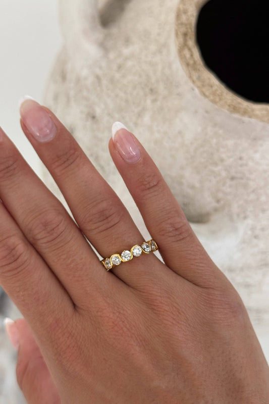 A close-up of a hand wearing a delicate gold band ring with five small clear round-cut stones, set against a soft-focus neutral background.