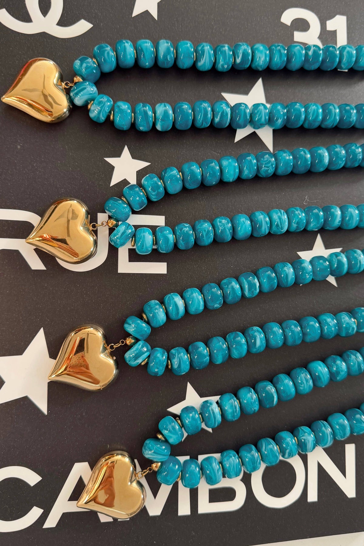 Four turquoise beaded necklaces with gold heart charms, arranged diagonally on a dark background with white star and text patterns.