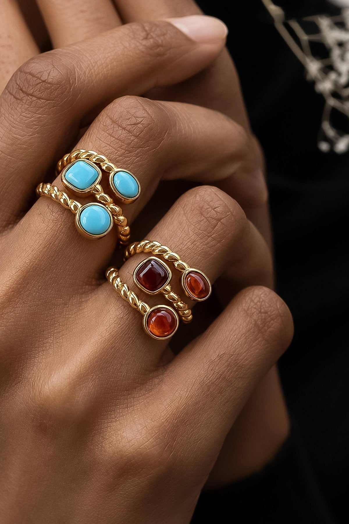 A close-up of a person's hand wearing three stacked gold rings with round turquoise, burgundy, and orange cabochon stones.