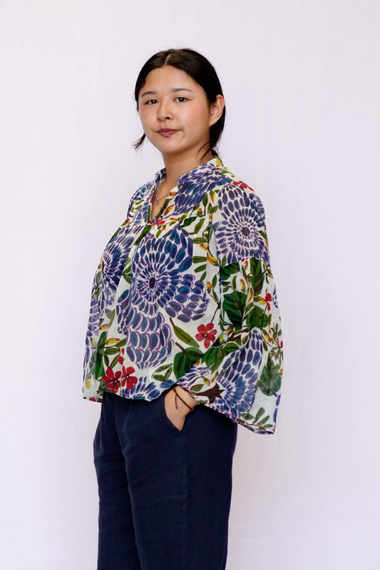 A woman wearing a long-sleeve button-up blouse with a vibrant blue, green, and red floral pattern on a white base, paired with dark navy pants, standing against a plain white background.
