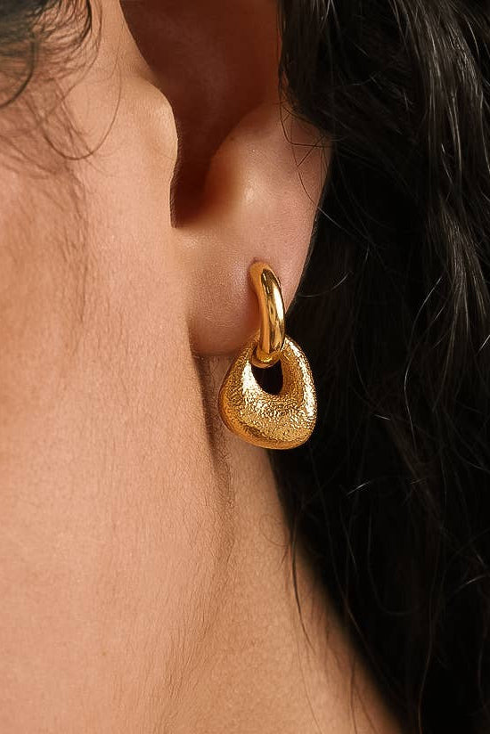A close-up of a textured gold huggie hoop earring worn on a person's ear, with dark hair and skin visible.