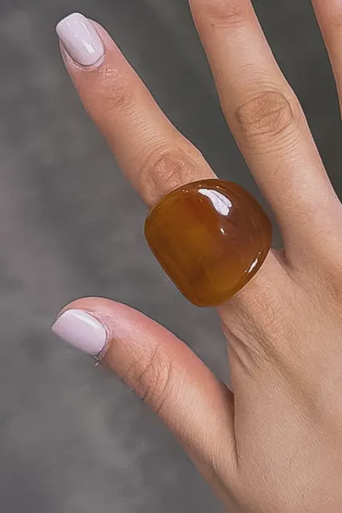 Hand wearing a large amber-colored ring on a neutral background