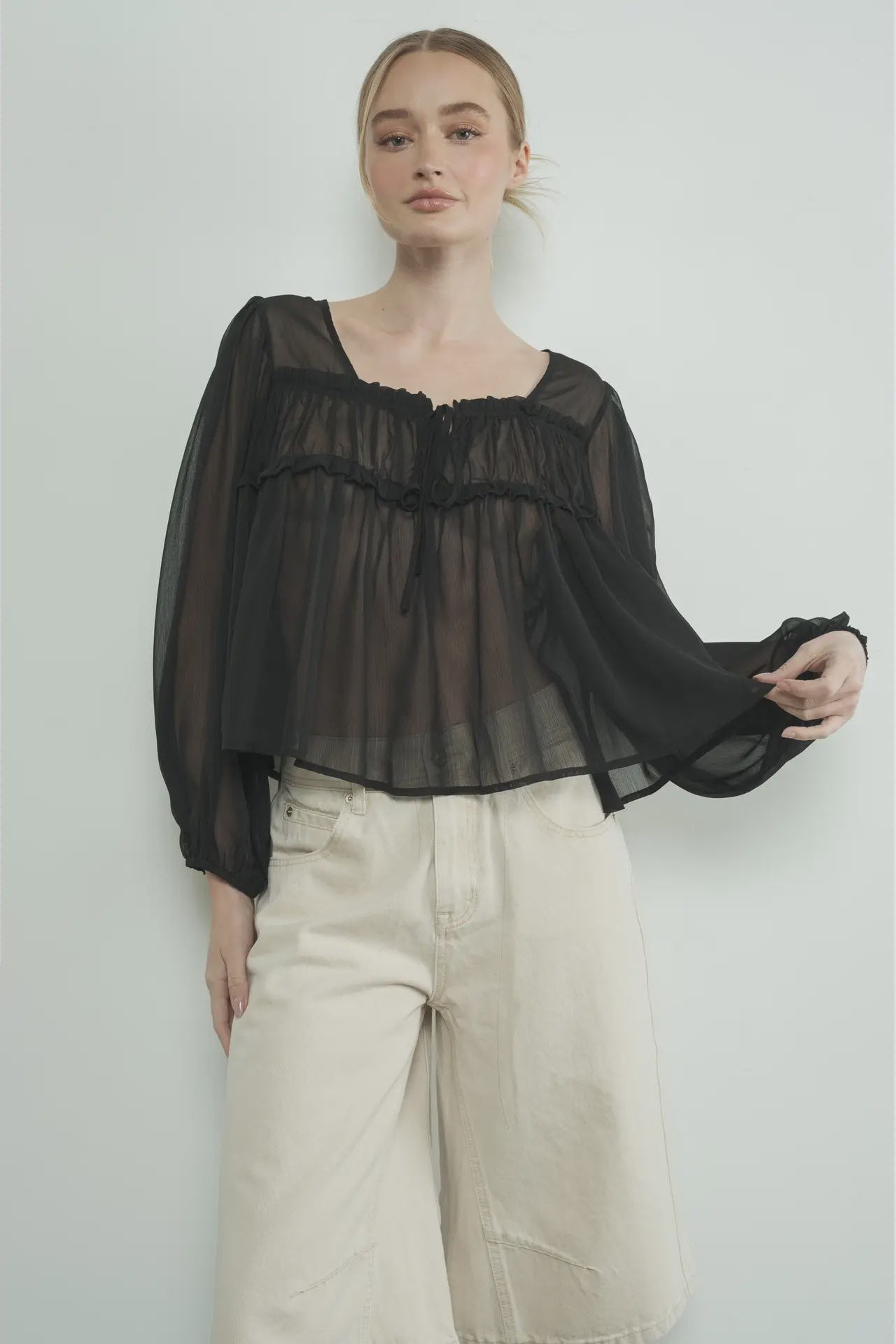 Person wearing a black sheer blouse and light-colored pants against a plain background