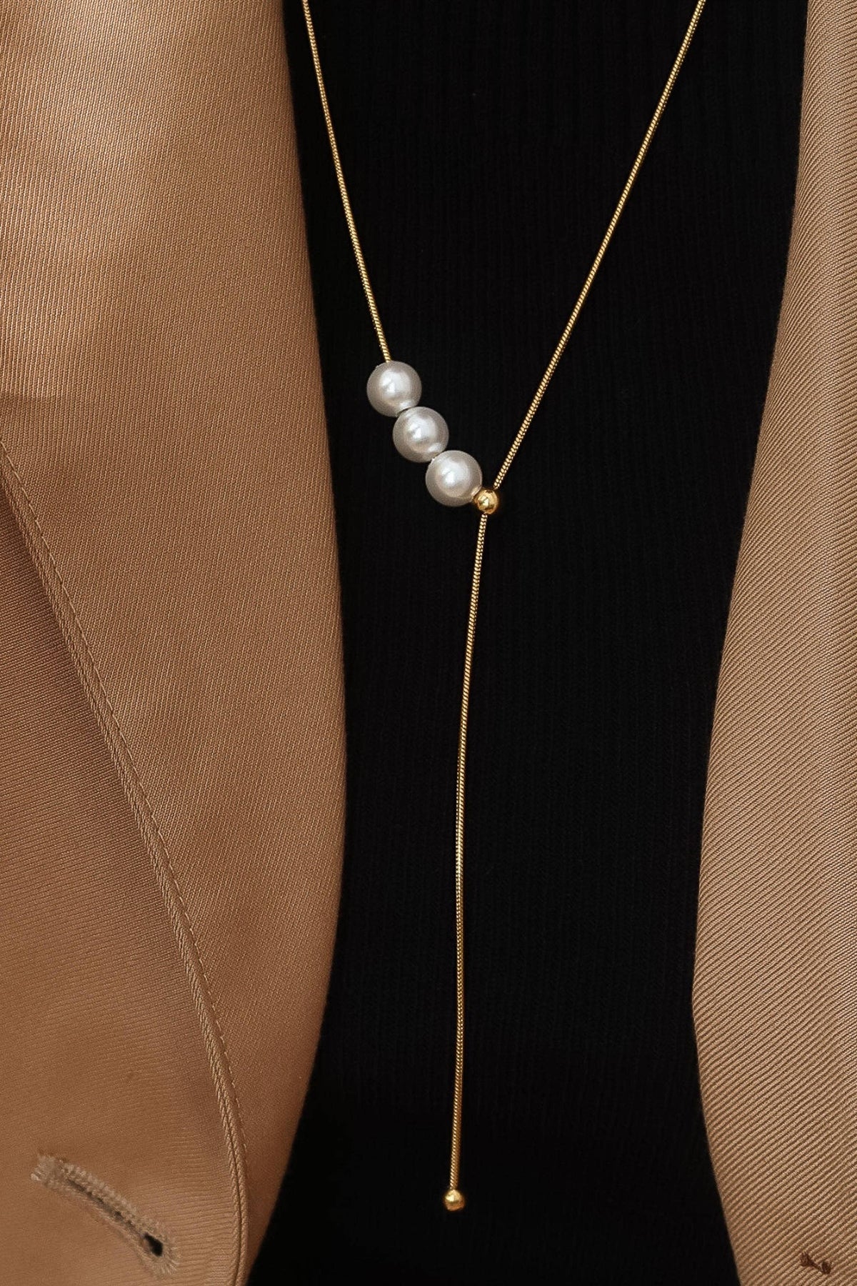 A gold Y-necklace with three white pearls, styled on a model wearing a black ribbed top and a tan blazer.