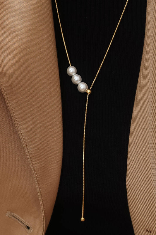 A gold Y-necklace with three white pearls, styled on a model wearing a black ribbed top and a tan blazer.