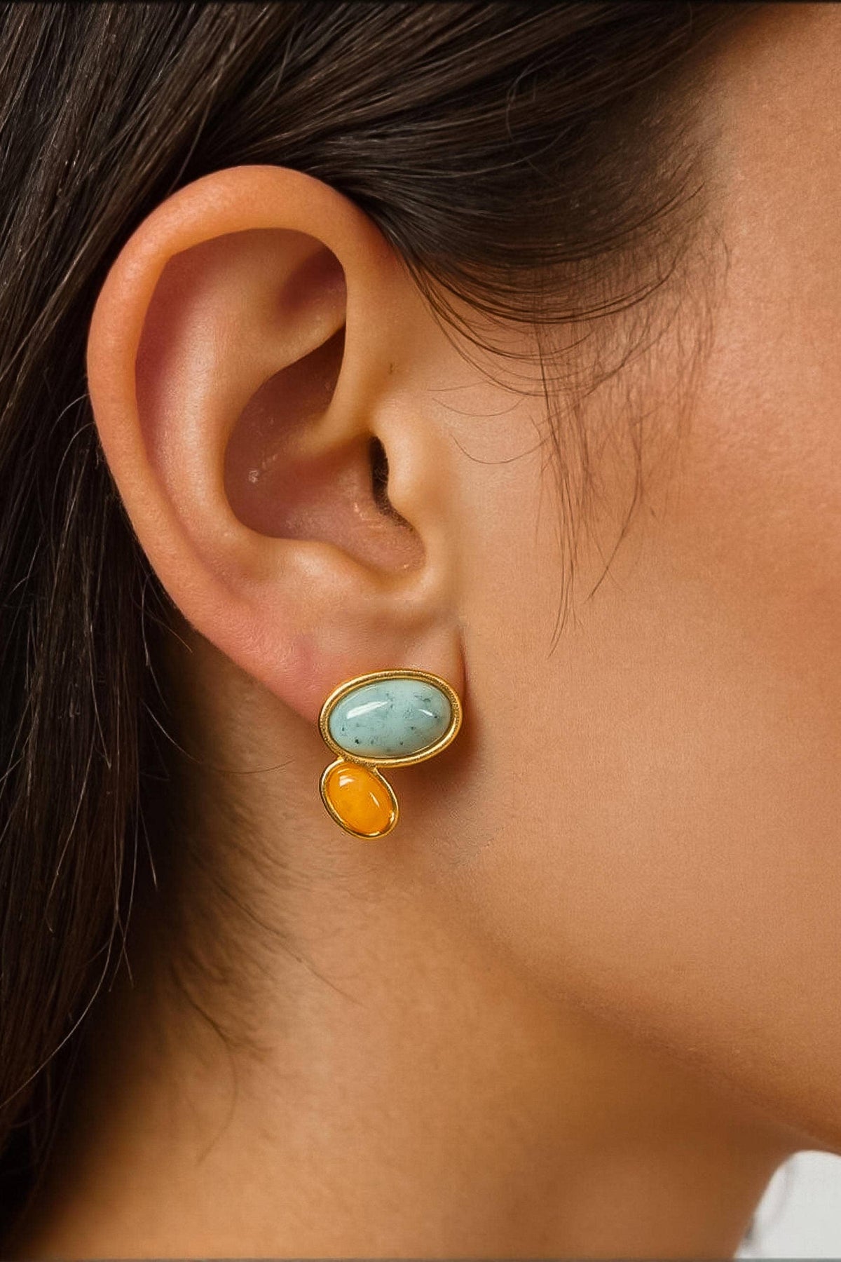 A close-up of a model's ear wearing a gold stud earring with a light blue oval stone and a small dangling gold and orange bead.