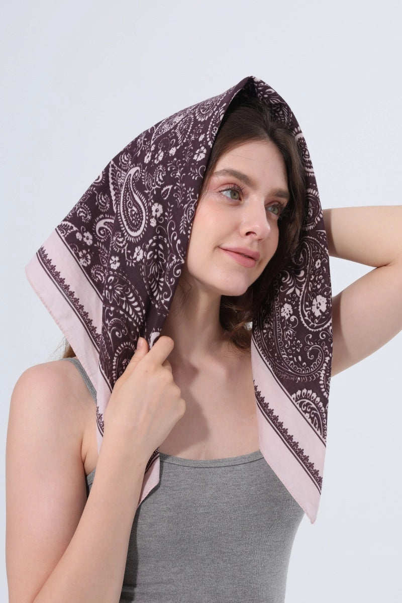 A woman wearing a dark brown paisley bandana with a light pink border, styled as a head covering. She is wearing a gray tank top against a plain light gray background.