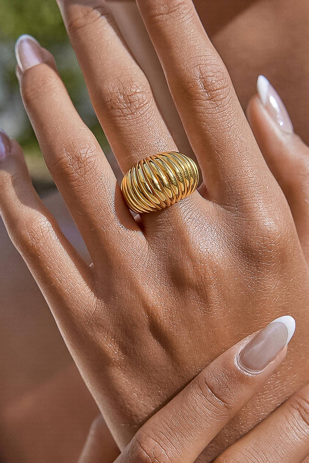 A close-up of a hand wearing a wide, ribbed gold ring with a polished finish. The hand has a warm medium-dark skin tone and manicured nails with a glossy nude finish.